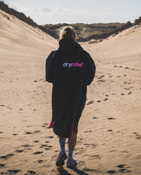 Woman walking away from camera through sand dunes, wearing Black Pink dryrobe® Advance Short Sleeve unzipped
