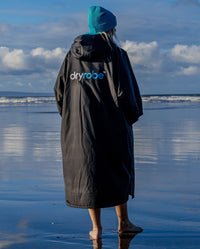 Woman walking towards the sea on a beach, wearing Black Blue dryrobe® Advance Long Sleeve