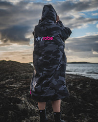 Woman stood on beach facing out to sea, wearing Black Camo Pink dryrobe® Advance Long Sleeve with hood up