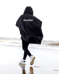 Girl running towards the sea on a beach, wearing Black Kids dryrobe® Waterproof Poncho
