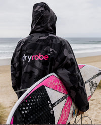 *MALE* facing the beach holding a surfboard, wearing Black Camo Pink dryrobe® Advance Long Sleeve