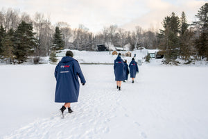 Back view of four people wearing custom Unbounded and Dryrobe® co-branded changing robes, walking through deep snow towards a winter campsite and cabins in the Canadian woods.