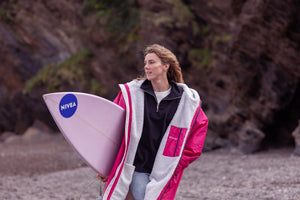 A woman wearing a bright pink dryrobe holding a surfboard on the beach