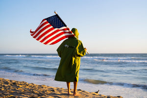 A person stood in a forest green dryrobe holding an American flag on the beach by the sea
