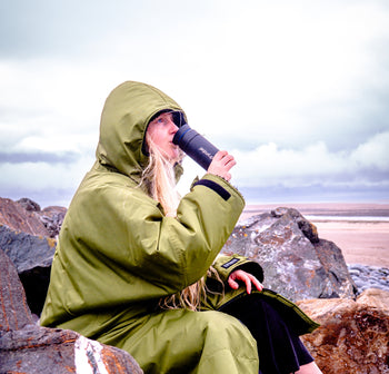 A girl sat on a rock at the beach drinking from a dryrobe branded Ocean Bottle wearing a dryrobe 