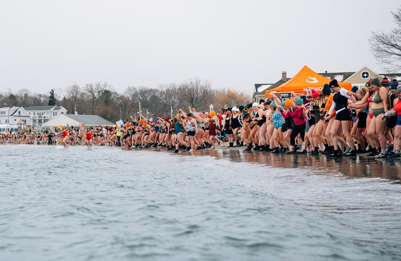 A wide shot of a massive crowd of women in swimwear and winter accessories lined up on a snowy beach, holding hands and preparing to charge into the cold water.