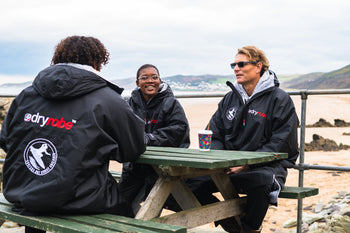 Tom Hewitt, Sne and Mini sat around a bench by the sea wearing Surfers Not Street Children dryrobes  