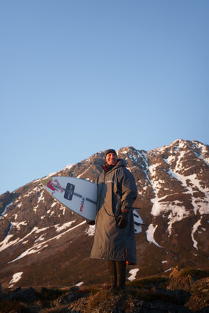 Andrew Cotton wearing a wetsuit and dryrobe holding a surfboard in Norway with snowy mountains behind him