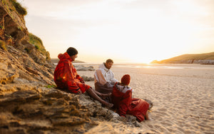Three people relaxing on rocky coastal terrain at the beach, staying warm in their dryrobes while watching the sun set over the water.
