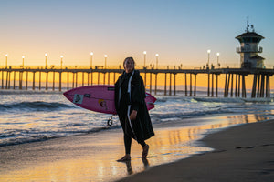 Izzi Gomez holding a surfboard wearing a wetsuit and dryrobe Lite on Huntington Beach, California 