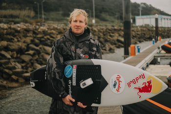 Big wave surfer Andrew Cotton in a dryrobe stood with his surfboard 