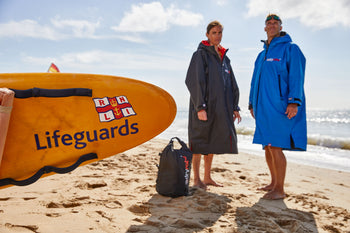 Two people in dryrobes stood next to a RNLI lifeguard board 
