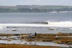Two surfers walking over rocks to the sea in Scotland wearing hooded wetsuits
