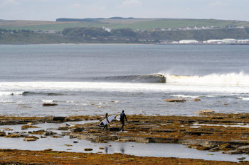 Two surfers walking over rocks to the sea in Scotland wearing hooded wetsuits
