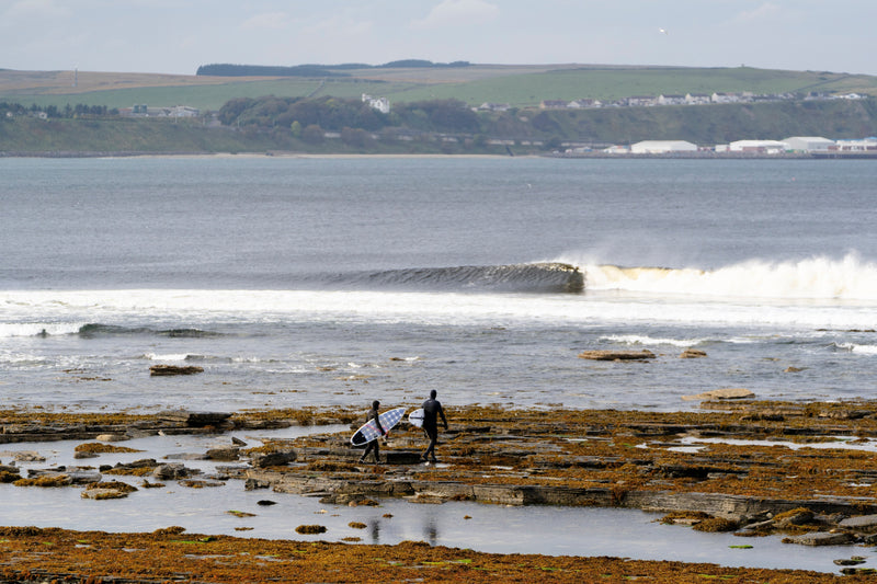 Two surfers walking over rocks to the sea in Scotland wearing hooded wetsuits