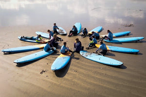 A group of young surfers and instructors sitting on their surfboards in a circle on the sand, engaging in a group discussion during a Liquid Therapy session.