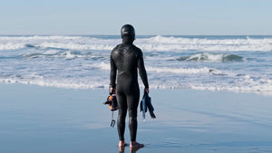 A surfer in a full black wetsuit and hood stands on a sunlit beach, looking out at the ocean waves while holding a camera housing and fins