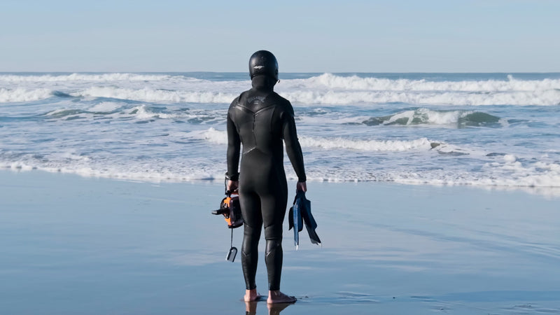 A surfer in a full black wetsuit and hood stands on a sunlit beach, looking out at the ocean waves while holding a camera housing and fins