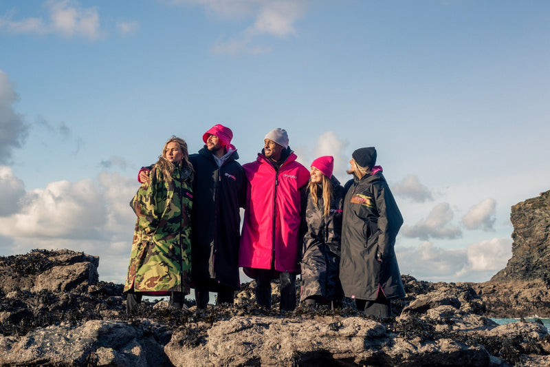 A group of people stood on a rock arm in arm looking out to sea and wearing dryrobes 