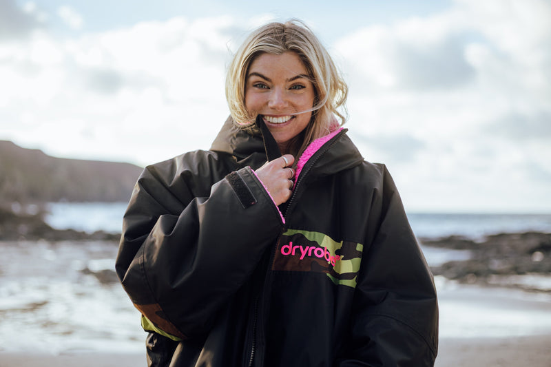A woman in a Dryrobe zipping it up and smiling at the camera while stood on the beach with the sea behind her 