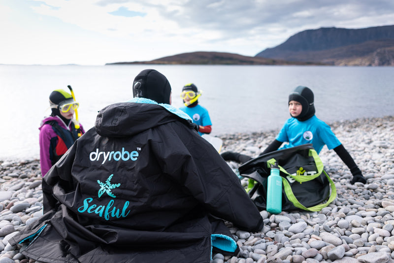 A group of children sat on a pebble beach wearing snorkelling gear with a Seaful branded Dryrobe wrapped around one of them