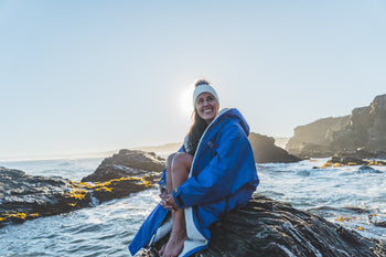 Bárbara Hernández Huerta  sat on a rock by the sea in a dryrobe® Advance change robe and swim hat 