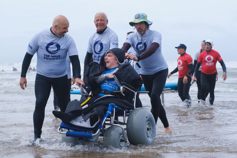 An adaptive surfer in a beach wheelchair wearing a Dryrobe Advance laughing while being assisted into the ocean by The Wave Project volunteers