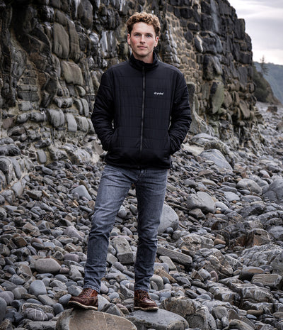 A man wearing a black quilted zip-up jacket, gray jeans, and brown boots stands on a rocky beach with a large stone cliff behind him. A small white logo is visible on the left chest of the jacket.