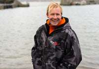 Person wearing a camouflage Dryrobe® changing robe with a brand logo standing by a body of water.