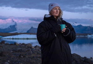 Person wearing a Dryrobe® changing robe holding a mug in a mountainous landscape with a lake.