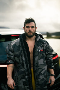 Man wearing a camouflage dryrobe with 'dryrobe' branding in front of a vehicle.