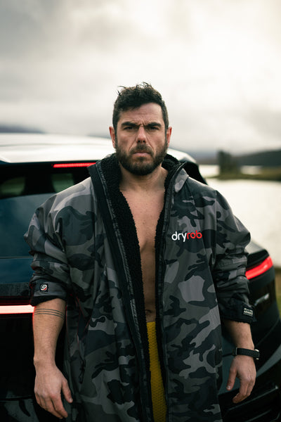 Man wearing a camouflage dryrobe with 'dryrobe' branding in front of a vehicle.
