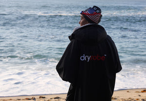 Person wearing a black Dryrobe® changing robe on a beach with ocean waves in the background