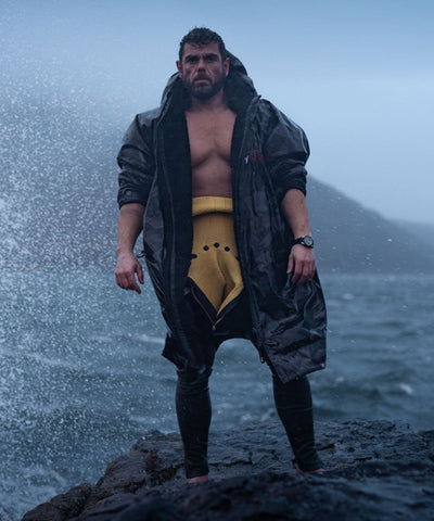Man in a Dryrobe® Changing robe and yellow wetsuit standing on a rocky outcrop with water splashing around.