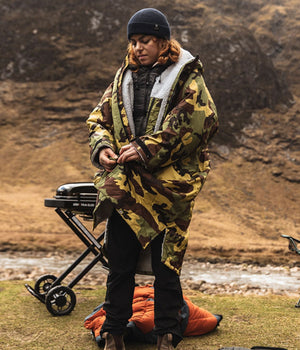 Person in camouflage grey Dryrobe® changing robe standing outdoors with a cart and orange bag in a natural setting
