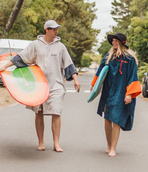 Two people wearing Dryrobe® full zip towelling changing robes holding surfboards on a road.