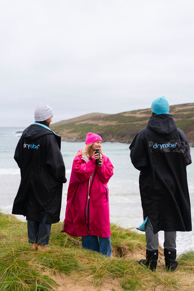 Three people wearing Dryrobe's standing on a grassy area by a body of water.
