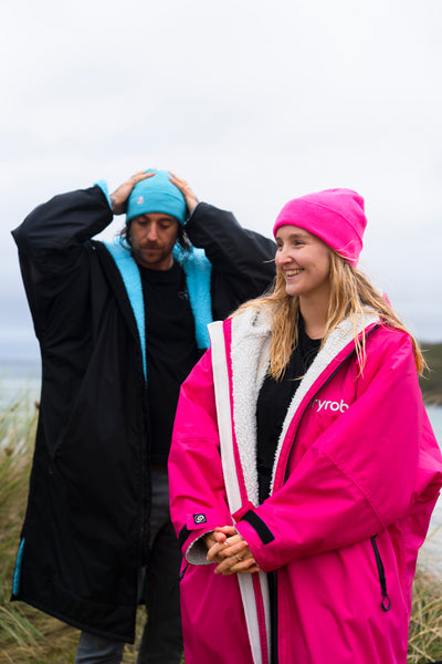 Two people wearing colorful Dryrobes and hats outdoors on a cloudy day.