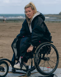 1|Woman sitting on wheelchair on a beach, wearing dryrobe® Adapt