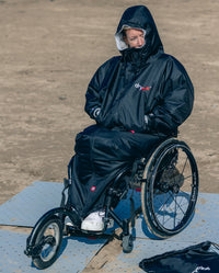 1|Woman sat on wheelchair on a beach, wearing dryrobe® Adapt with the hood up 