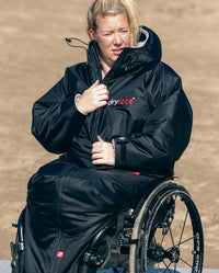 1|Woman sat on wheelchair on a beach, wearing dryrobe® Adapt with the hood down