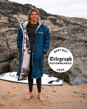 A man stands barefoot on a sandy beach in front of rocky cliffs, wearing a long navy blue Dryog waterproof jacket over a black shirt and black leggings. A white surfboard lies on the sand behind him, and a circular badge on the right reads 'Best Buy Telegraph Recommended 2026'.