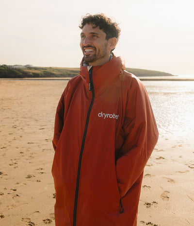 *MALE* A man smiling on a sandy beach wearing a red-orange Dryrobe waterproof Changing robe, The background shows water and distant land under a bright sky.