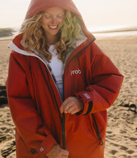 A woman wearing a bright red waterproof hooded Changing robe, with a white inner lining stands on a sandy beach. The jacket has a white 'yrobo' logo on the left chest and a white brand patch on the cuff.