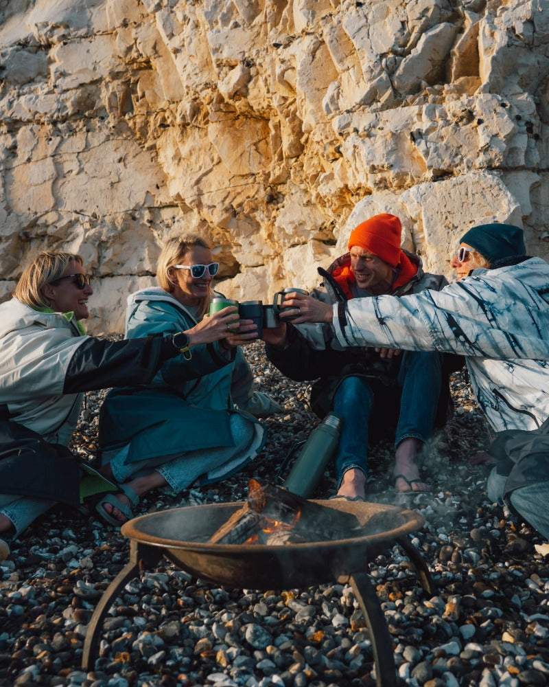 Four people sitting on a pebble beach in front of a large rock face, clinking cups together around a small fire pit. They are wearing outdoor Dryrobe's and beanies in warm natural light.