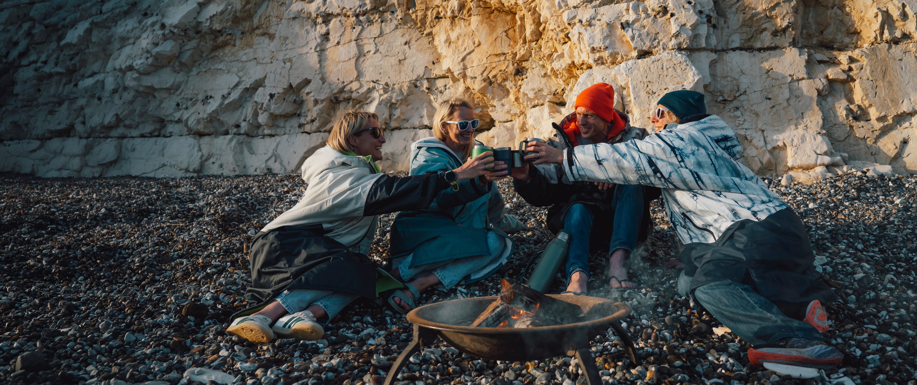 Four people sitting around a small fire pit on a pebble beach in front of a large rock face, sharing a moment with warm drinks. They are dressed in casual outdoor clothing, with one person wearing a bright orange beanie.
