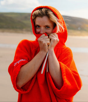 A woman on a beach wearing a bright orange hooded poncho, holding the hood up over her face with both hands. The brand name 'dryrobe' is visible in white text on the left sleeve.