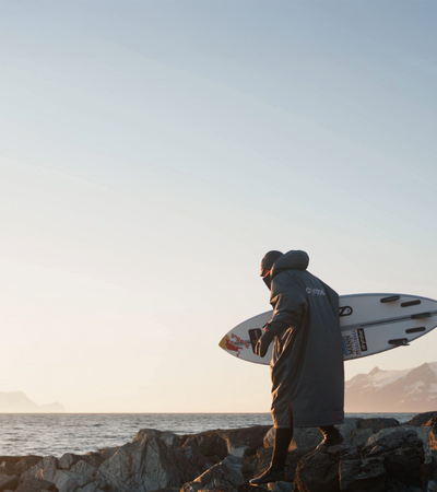 Person holding a surfboard on a rocky shoreline with mountains in the background
