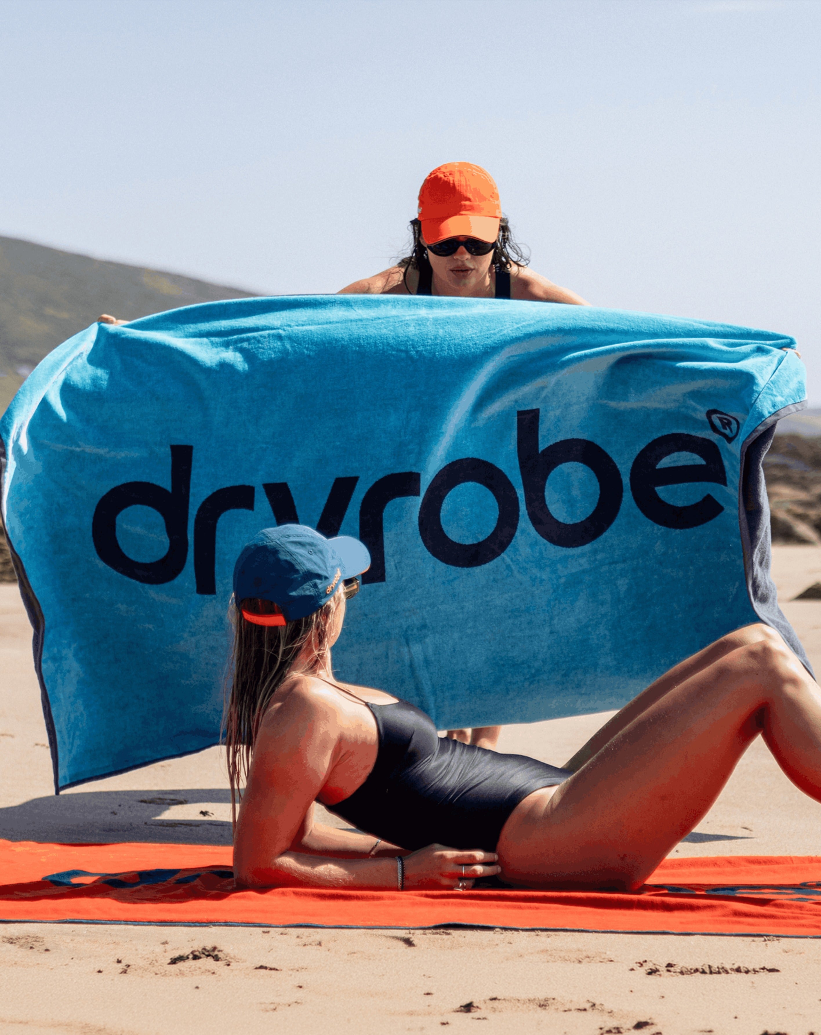 A woman in a black swimsuit lies on a red towel on a sandy beach while another woman holds up a large blue Dryrobe towel behind her. The background shows a beach and distant hills under a clear sky.