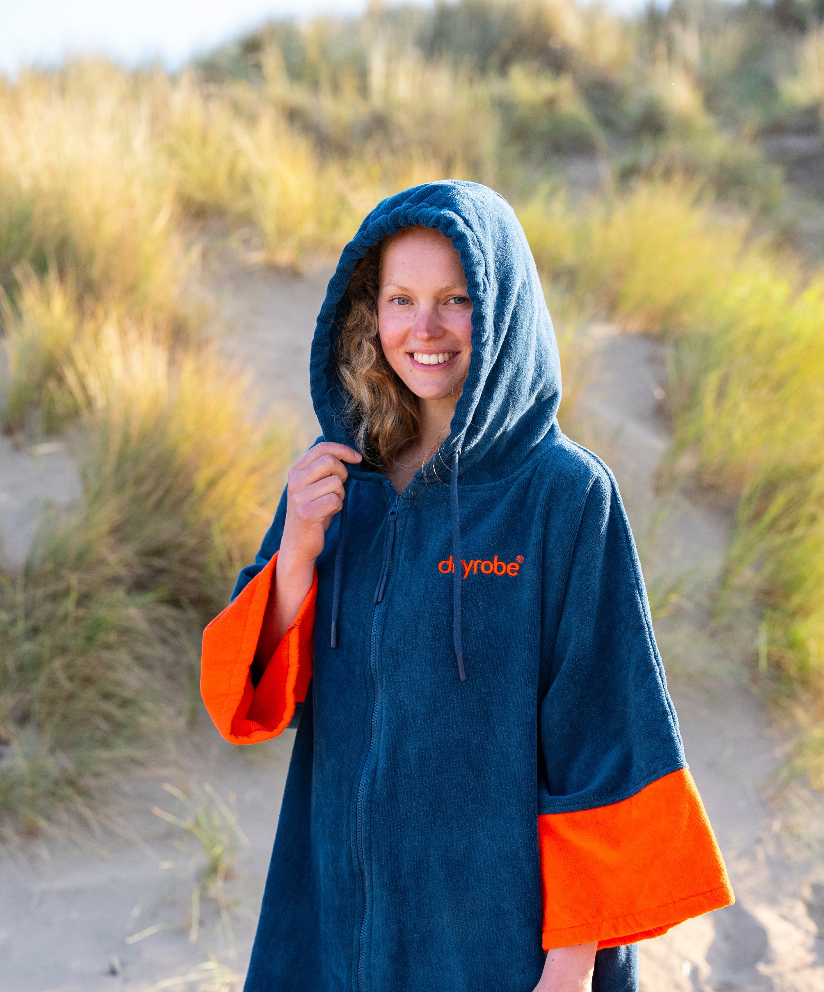 A woman wearing a navy blue hooded robe with bright orange cuffs and hem, standing on a sandy path in a grassy dune landscape. The brand name 'dryrobe' is printed in orange on the chest.